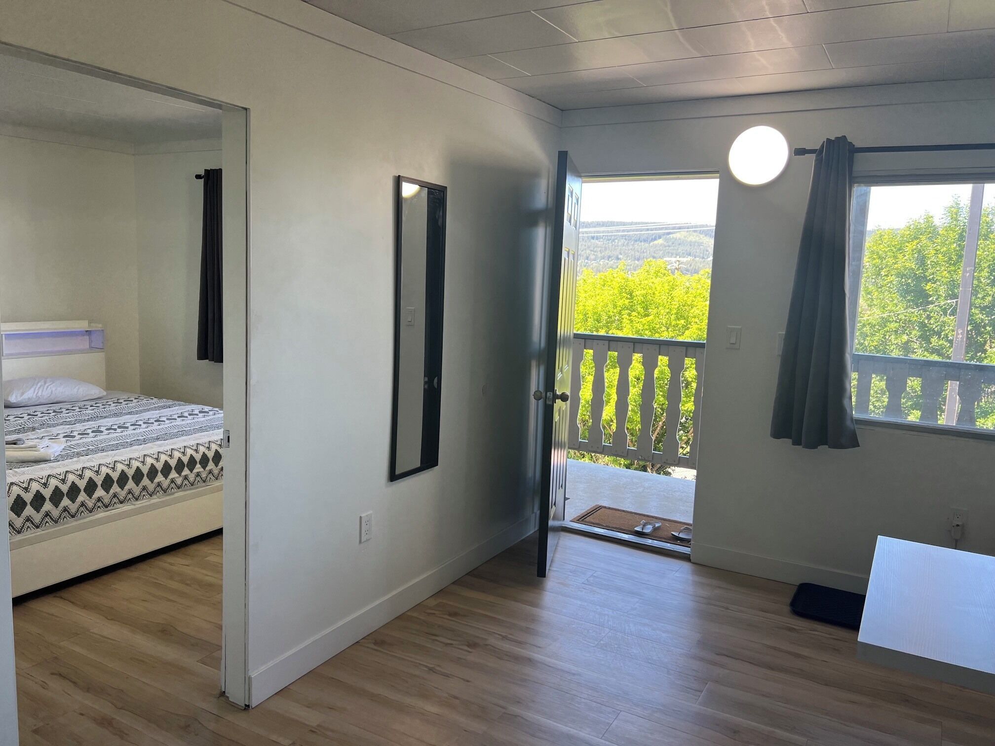 A bedroom with a bed, a mirror, a door leading to a balcony, and a view of greenery.