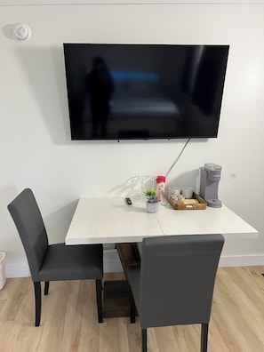 Un comedor con una mesa blanca, dos sillas negras, un televisor de pantalla plana montado en la pared y una cafetera sobre la mesa.