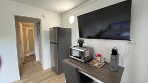 Una kitchenette compacta con refrigerador, microondas y cafetera, una mesita con una silla y un televisor montado en la pared.