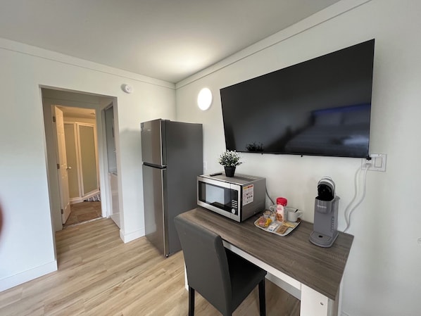 Una kitchenette compacta con refrigerador, microondas y cafetera, una mesita con una silla y un televisor montado en la pared.