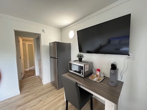 A compact kitchenette with a refrigerator, microwave, and coffee maker, a small table with a chair, and a TV mounted on the wall.