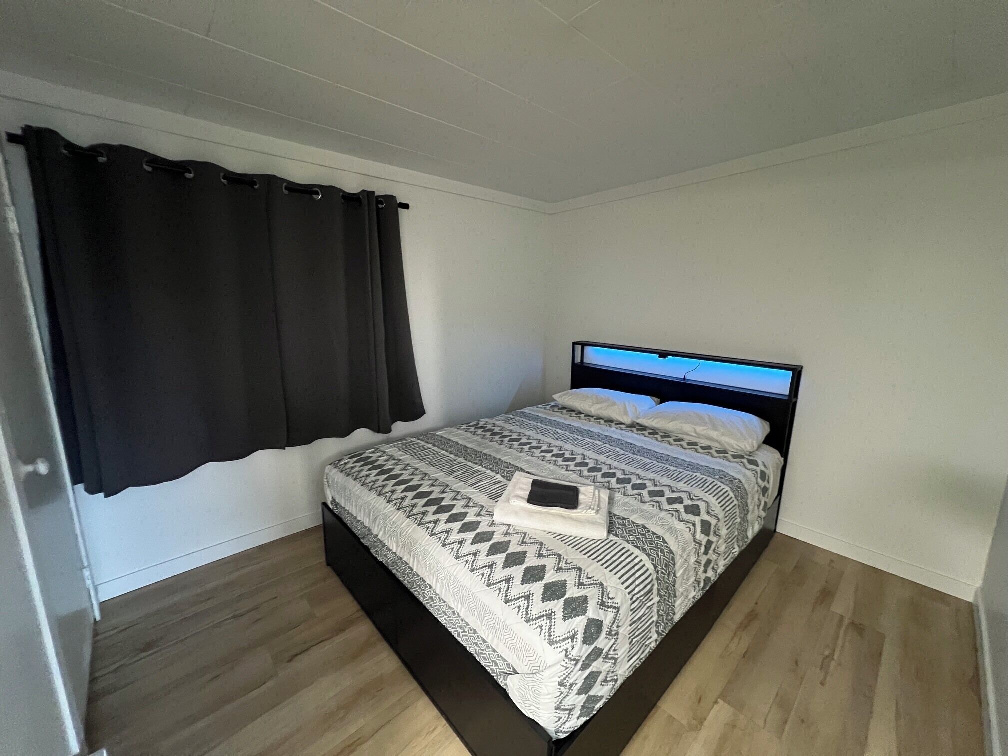 A single bed with a patterned bedspread, a black headboard, and a window with black curtains.