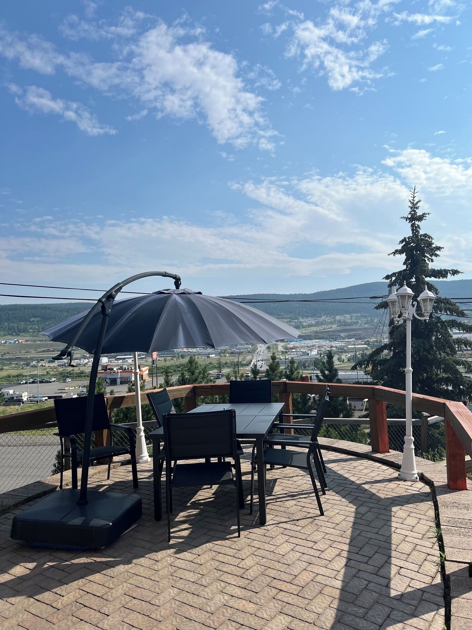 An outdoor seating area with a table and chairs, a large umbrella, and a view of a landscape with trees and buildings.