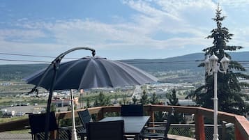 An outdoor seating area with a table and chairs, a large umbrella, and a view of a landscape with trees and buildings.