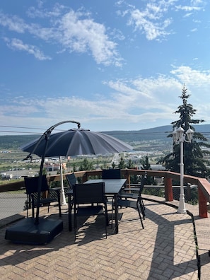 An outdoor seating area with a table and chairs, a large umbrella, and a view of a landscape with trees and buildings. - Stampeder Inn (Williams Lake)