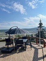 Un espacio al aire libre con una mesa y sillas, un paraguas grande y vistas a un paisaje con árboles y edificios.
