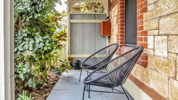 A covered outdoor seating area with two black wire chairs, a stone wall, and a plant-covered trellis. - Ada Art Deco by Host Solutions Hutt St, Heritage (Adelaide)