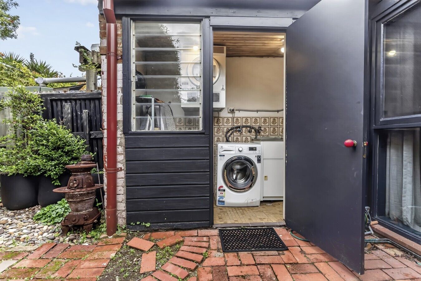 A laundry room with a washing machine, a drying rack, and a door leading to a garden area with potted plants and a brick pathway.