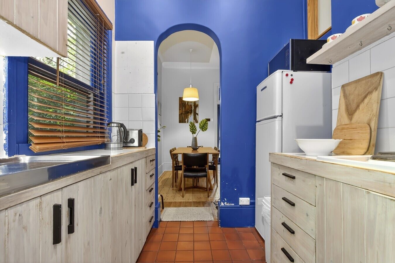 A narrow kitchen with blue walls, a window with blinds, a dining area with a table and chairs, and a refrigerator.