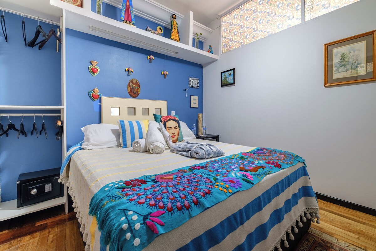 A bedroom with a bed, a nightstand, a wall-mounted shelf with decorative items, and a window with patterned curtains.