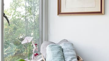 Una habitación con un ventanal grande, una cesta de mimbre con toallas, un pequeño jarrón con una planta y un cuadro en la pared.