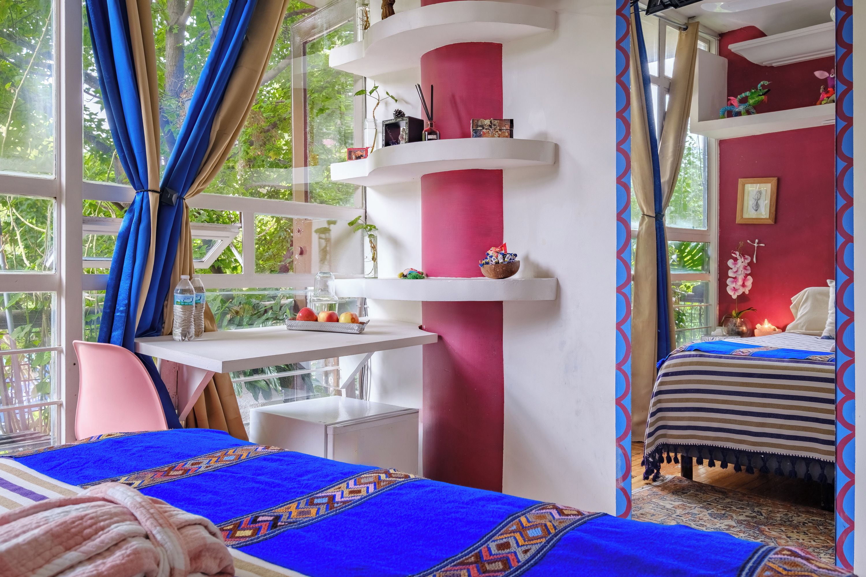 Un dormitorio con cama, escritorio y silla. Hay estanterías con objetos decorativos y una ventana con cortinas.
