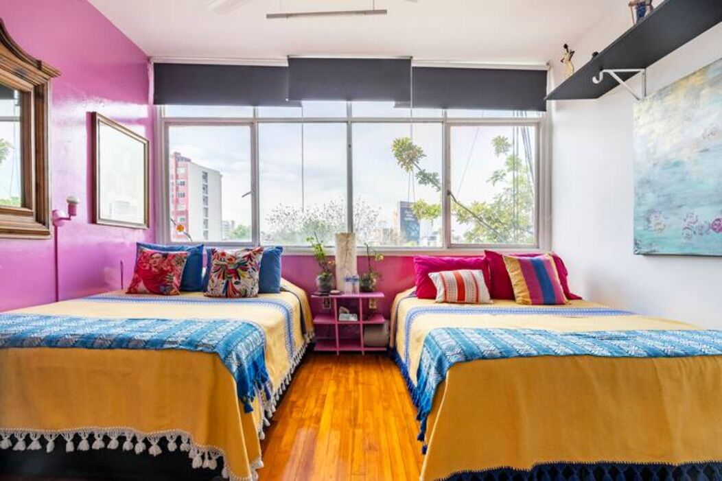 Una habitación con dos camas, ventanas amplias, una pared rosa y un cuadro.