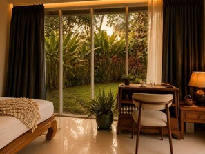 Ein Zimmer mit einem Bett, einem Schreibtisch, einem Stuhl, einem Tisch mit Lampe und Blick auf einen Garten durch große Fenster.
