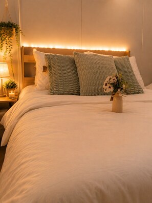 Ein ordentlich bezogenes Bett mit weißer Decke und zwei grünen Kissen, ein Nachttisch mit Lampe und einer kleinen Pflanze sowie eine wandmontierte Leuchte.