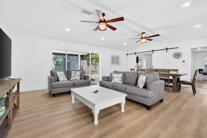 Ein modernes Wohnzimmer mit zwei grauen Sofas, einem weißen Couchtisch, Deckenventilatoren und einem Essbereich mit einem Holztisch und Stühlen.