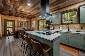 A modern kitchen with a central island, bar stools, and a view of the forest through large windows. - Timber Vail | Luxury Cabin in the Woods (Sevierville)