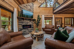 A cozy interior with leather sofas, a stone fireplace, and a view of the outdoors. - Timber Vail | Luxury Cabin in the Woods (Sevierville)