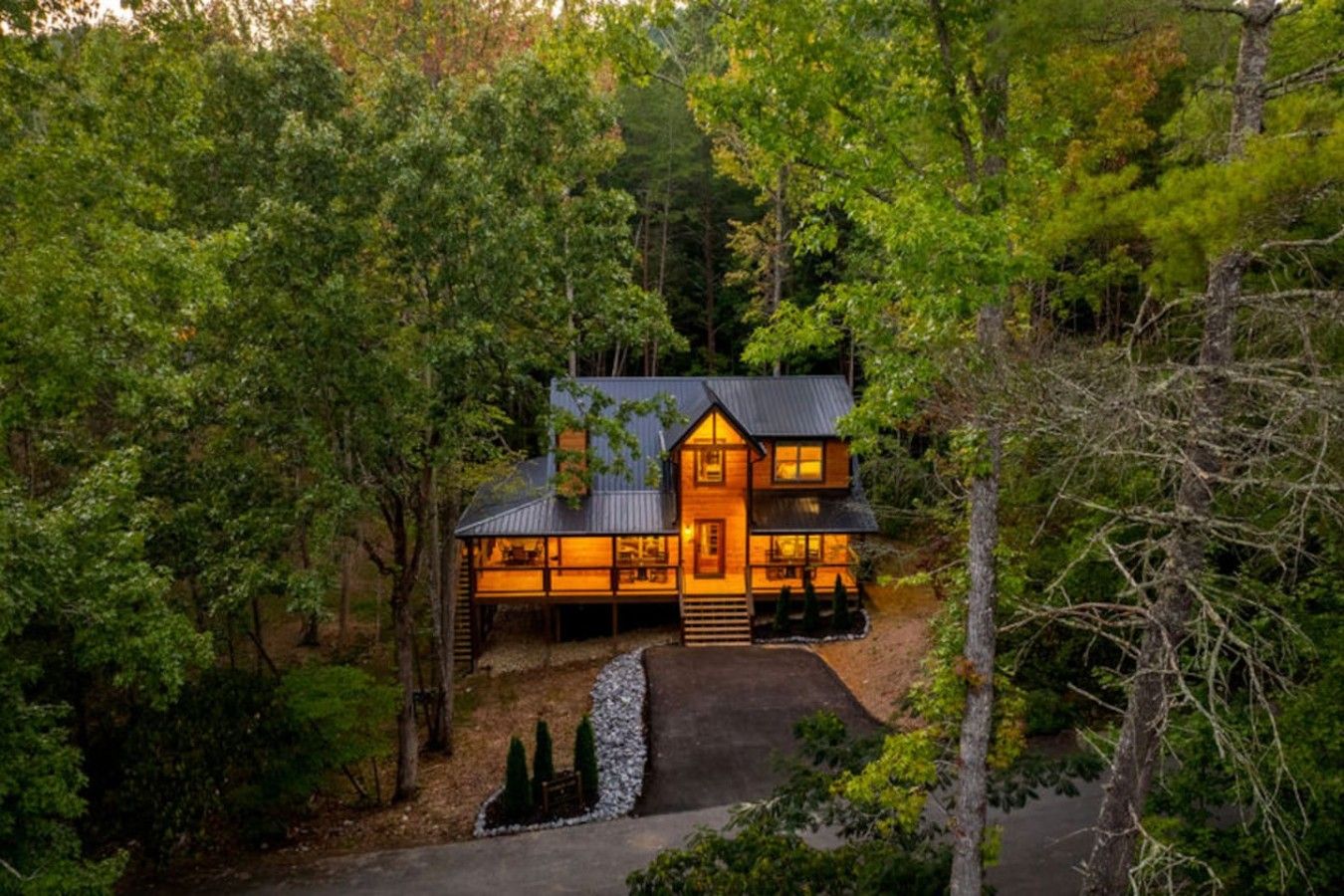 A two-story wooden cabin surrounded by dense forest, with a visible front porch and a pathway leading to the entrance.
