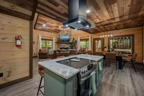 A spacious kitchen with a central island, wooden walls, and a stone fireplace. - Timber Vail | Luxury Cabin in the Woods (Sevierville)