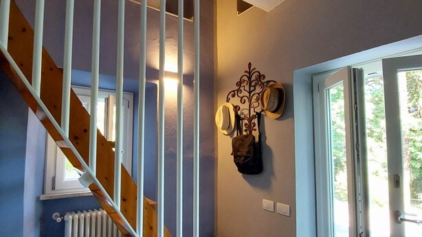 A staircase with wooden railings, a wall-mounted hat rack, a radiator, and a door leading to a bright room with a view of greenery. - Acquazzurra, luxury duplex, pool, parking space, free WiFi, By Guestaly (Lesa)