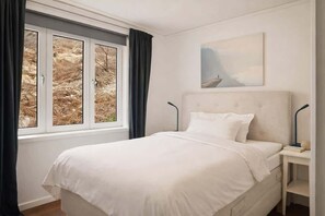 Et soveværelse med en seng, et natbord, en lampe, et vindue med gardiner og et billede på væggen.
