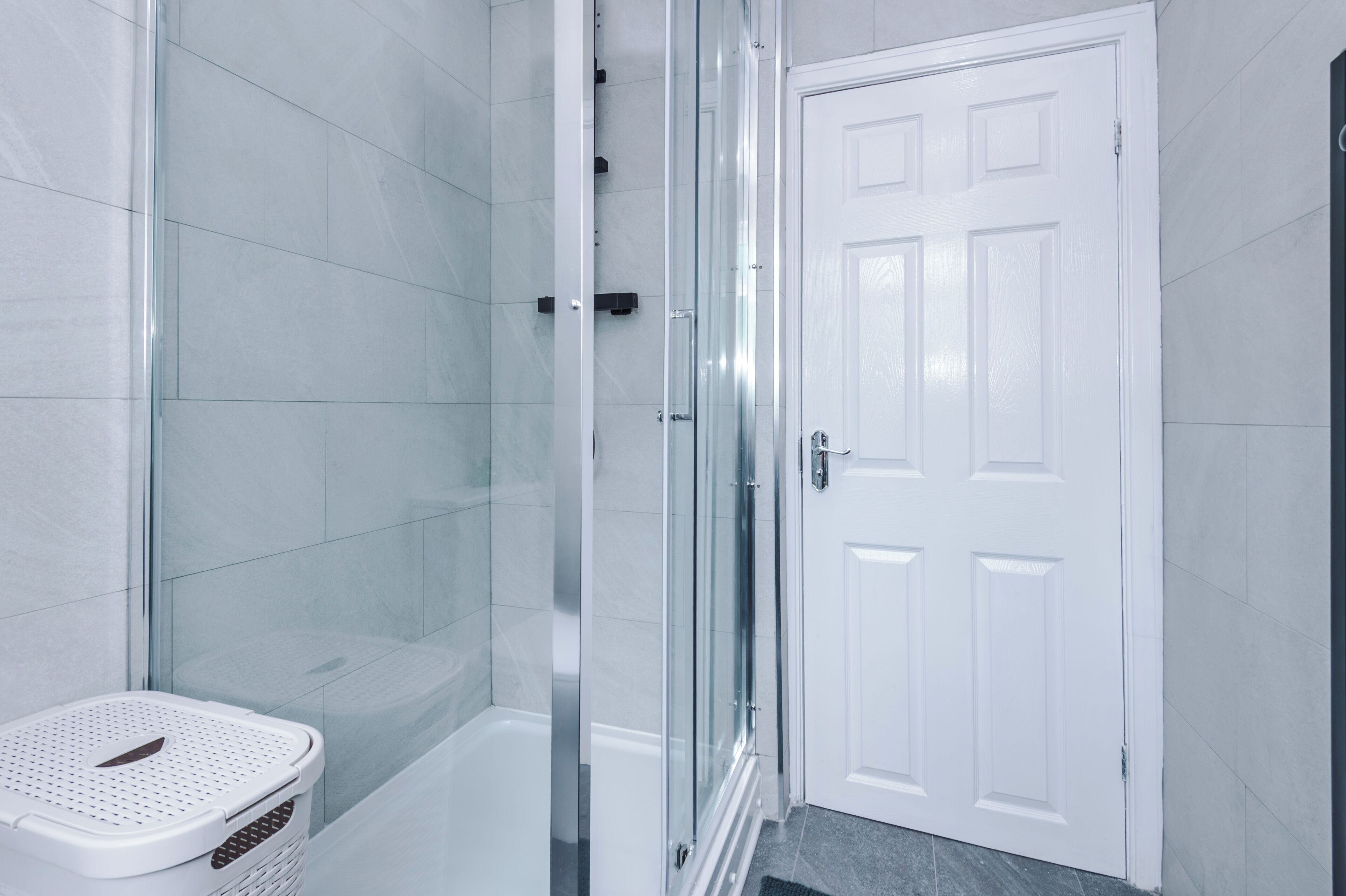 A bathroom with a glass shower enclosure, a white door, and a white laundry basket.