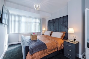 A modern bedroom with a bed, bedside lamps, a television, and a window with blinds. - 2 Bed House with Driveway & Workspace  (Manchester)