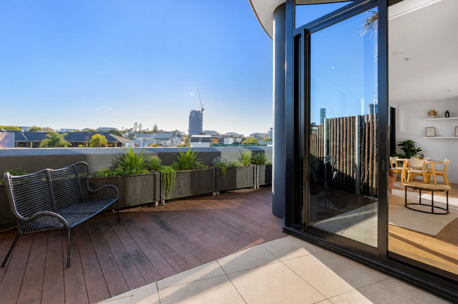 Un balcon moderne doté d’un plancher en bois, d’un banc en métal et d’une porte vitrée donnant sur un espace intérieur comprenant un coin repas.