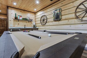 A pool table with a cue stick and a billiard ball, wooden walls with a shelf holding bottles, and a wall-mounted wheel. - Aska Timber Lodge | Blue Ridge, GA (Cherry Log)