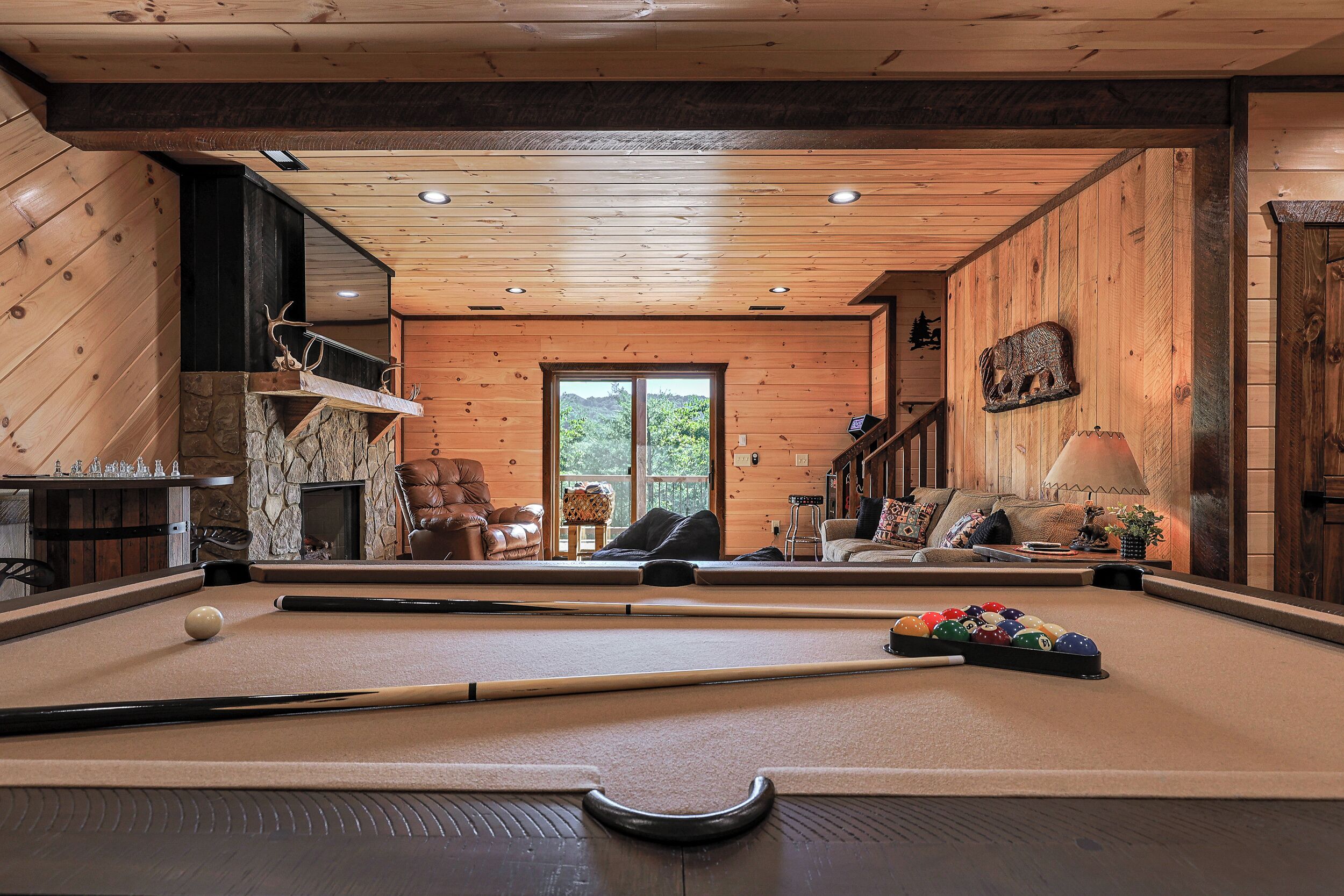 A pool table in a wooden room with a fireplace, seating area, and a view through a glass door.