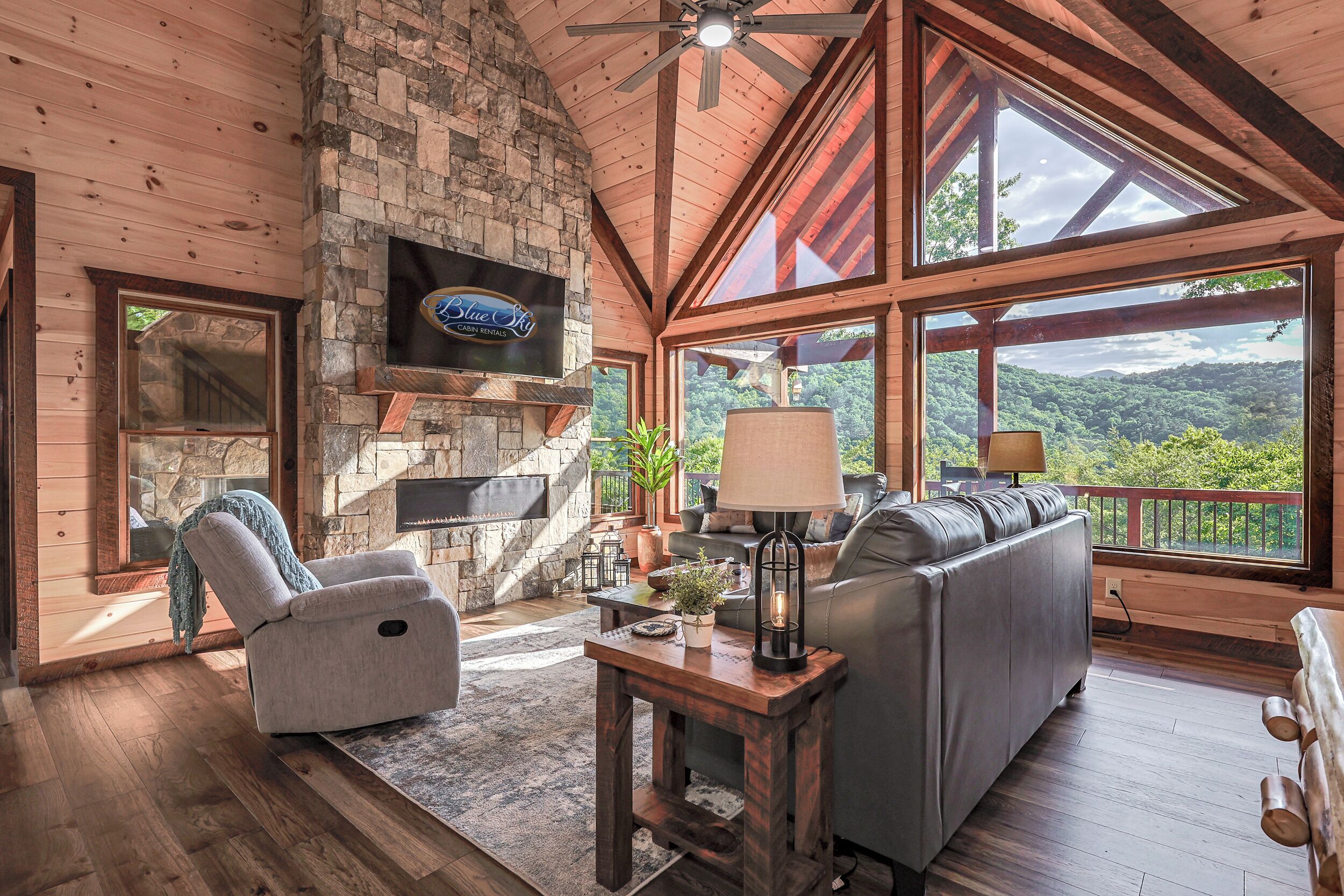 A spacious living area with a stone fireplace, a flat-screen TV mounted on the wall, a comfortable sofa, and large windows offering a view of the outdoors.