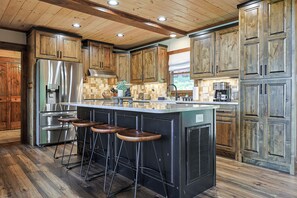 A kitchen with wooden cabinets, a stainless steel refrigerator, a large island with stools, and a window with a view. - Aska Timber Lodge | Blue Ridge, GA (Cherry Log)