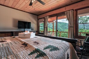 A bedroom with a large bed, a flat-screen TV, a view of a forested area, and a balcony with a chair. - Aska Timber Lodge | Blue Ridge, GA (Cherry Log)