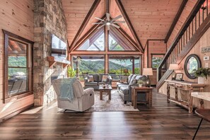 A spacious living area with a stone wall, wooden ceiling, and large windows offering a view of the outdoors. - Aska Timber Lodge | Blue Ridge, GA (Cherry Log)