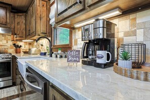 A kitchen with wooden cabinets, a marble countertop, a coffee maker, and a sign that reads 'Coffee keeps Mr. going.' - Aska Timber Lodge | Blue Ridge, GA (Cherry Log)