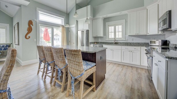 A kitchen with white cabinets, a central island with bar stools, a hanging light fixture, and a fish decoration on the ceiling. - 1327 - Seaglass Retreat, Corolla Retreat | Hot Tub, Pool & 4BR Comfort (Corolla)
