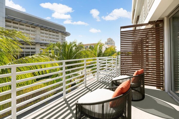 Ein Balkon mit weißer Reling, zwei schwarze Korbstühle mit Kissen und Blick auf ein Gebäude mit Kuppel und Palmen.
