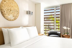 Ein Hotelzimmer mit einem großen Bett, einem Wandbild, einem Fenster mit Jalousien, einem Sessel und einem kleinen Tisch mit einer Pflanze.