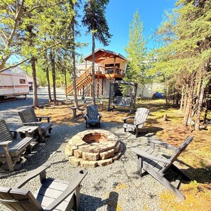 En campingplats med en eldstad, trästolar och en stuga i bakgrunden.