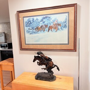 Pintura emoldurada de uma paisagem nevada com cavalos e uma escultura de um cavaleiro montado sobre uma mesa de madeira.