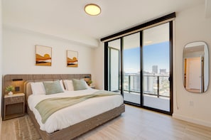 A modern bedroom with a large bed, a nightstand, a mirror, and a view of the city through floor-to-ceiling windows. - AEA - Carlyle Avenue FL (Miami Beach)