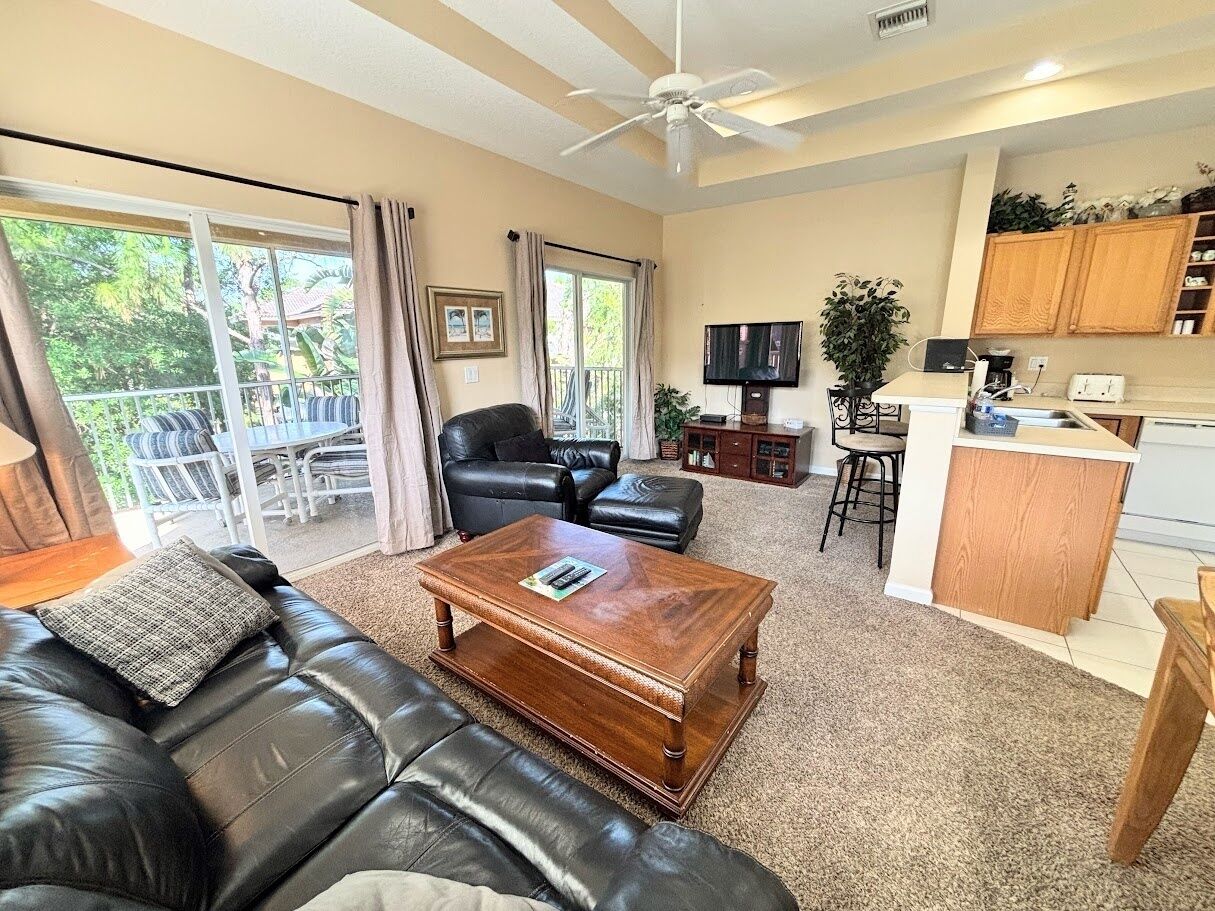 A living room with a leather sofa, a wooden coffee table, a TV, and a kitchen area with a counter and cabinets.