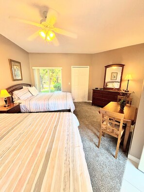 A bedroom with two beds, a ceiling fan, a desk with a chair, a dresser, and a window with a view of greenery. - Right Direction Rentals Studio Villa 8249 (Port St Lucie)