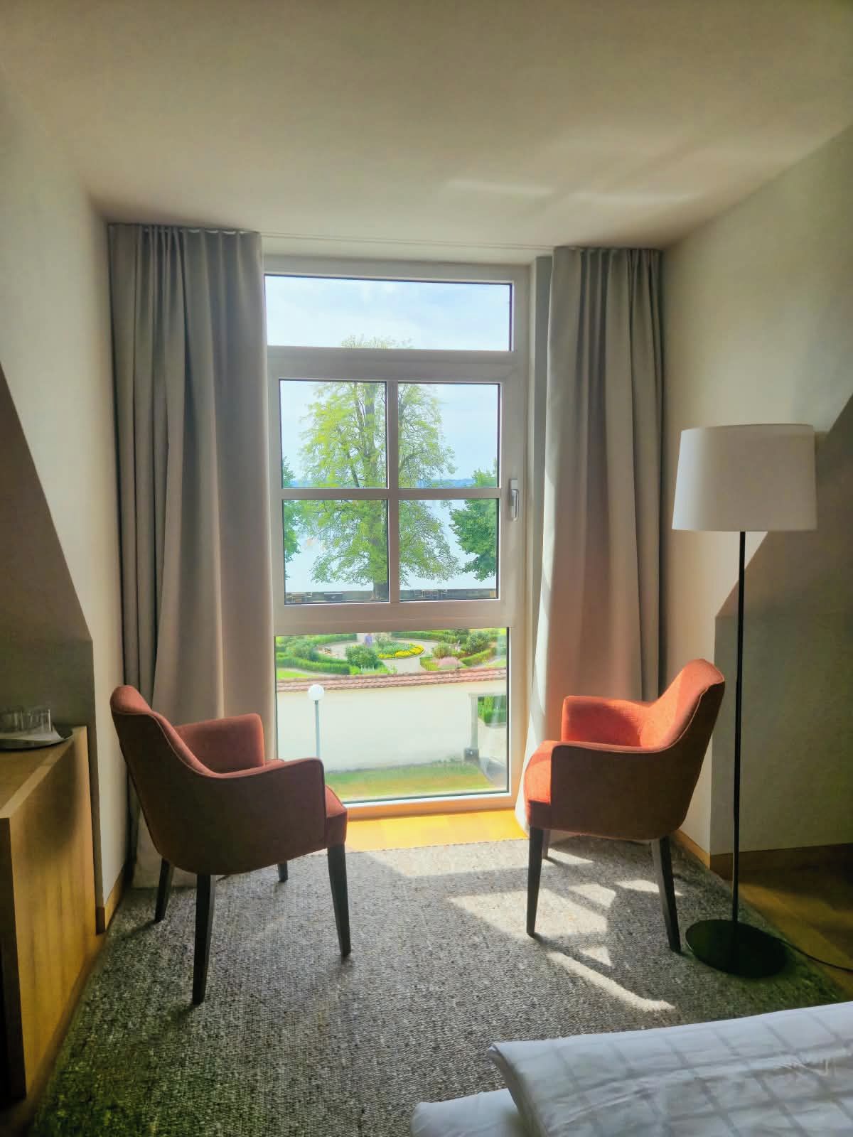 Quarto de hotel com uma grande janela, duas poltronas, uma mesa de cabeceira, um abajur e uma cama.