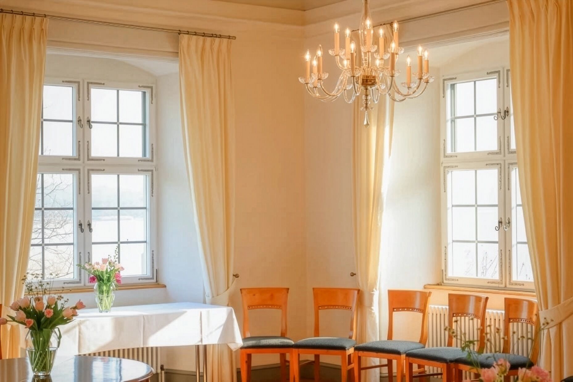 Un espace repas comprenant une table, des chaises, un lustre et de grandes fenêtres avec des rideaux.