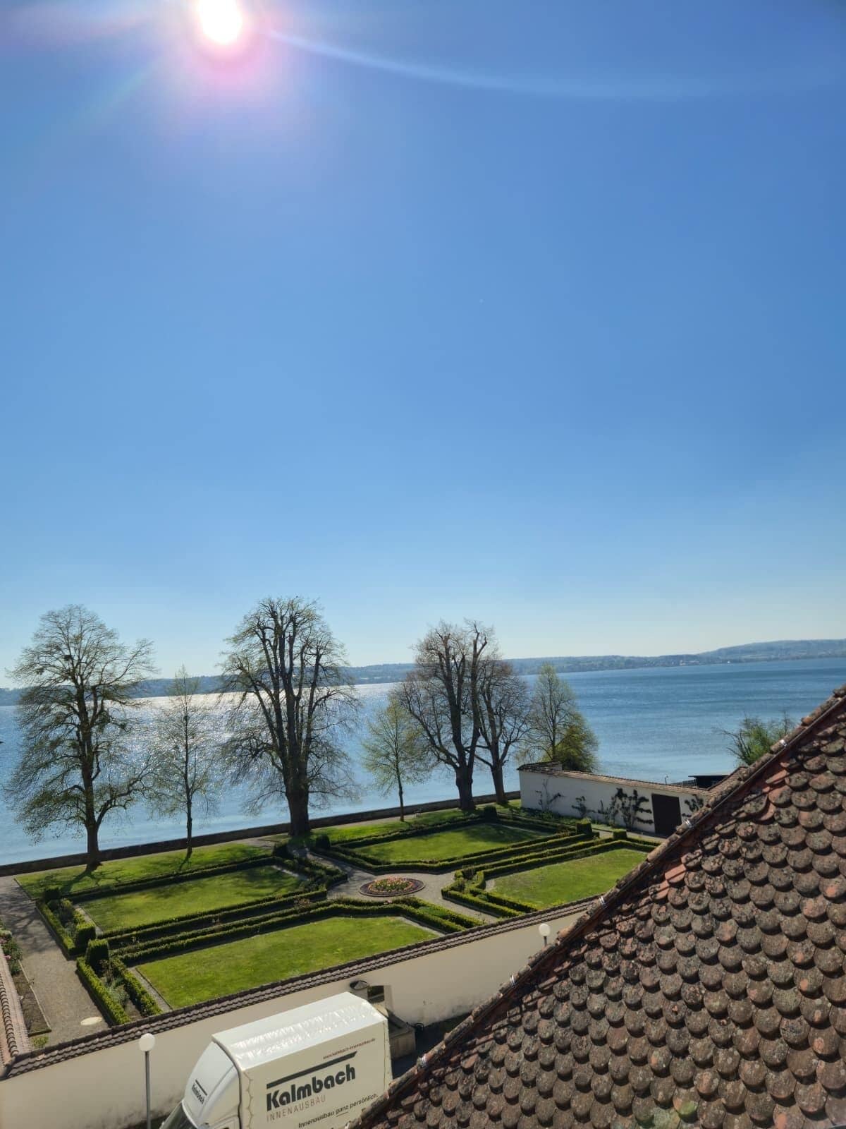Vista de un jardín ajardinado con un cuerpo de agua al fondo, cielo despejado y un edificio con techo visible.