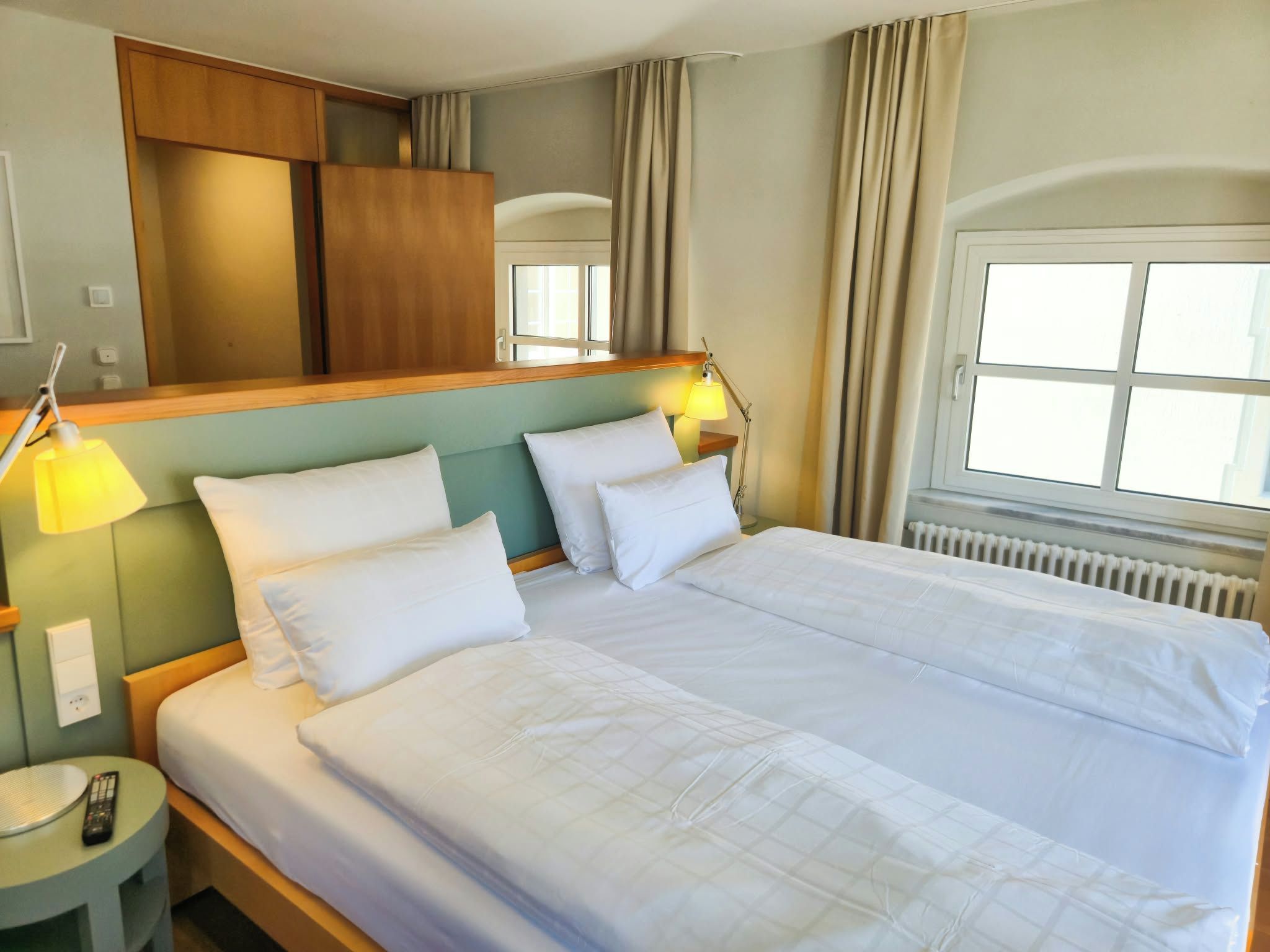 Habitación de hotel con cama doble, lámparas de noche, mesita de noche, espejo y ventana con cortinas.