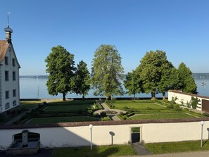 A serene lakeside view with a well-maintained garden, a white building with a tower, and a clear sky.