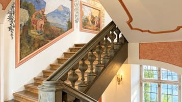 Eine prächtige Treppe mit aufwendig gestalteten Geländern und einer Handlauf, die zu einer Zwischenebene mit einem großen Wandgemälde und einem Fenster mit Blick auf Grünflächen führt.